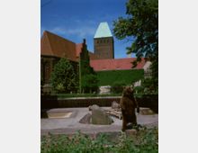 Ein brauner Bär steht aufrecht in einem umzäunten Gehege mit Steinen und Baumstämmen. Im Hintergrund sind ein rotes Ziegeldach, eine mit Efeu bewachsene Gebäudeaußenwand und ein Turm mit grünem Kupferdach sichtbar, umgeben von Bäumen.