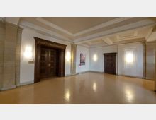 Geräumiger Flur in einem Gebäude mit heller Wandgestaltung, Stuckverzierungen und zwei hölzernen Türen, von denen eine mit der Aufschrift "Magistratssitzungszimmer" versehen ist; eine Wand enthält ein gerahmtes Bild und ein Hinweisschild.