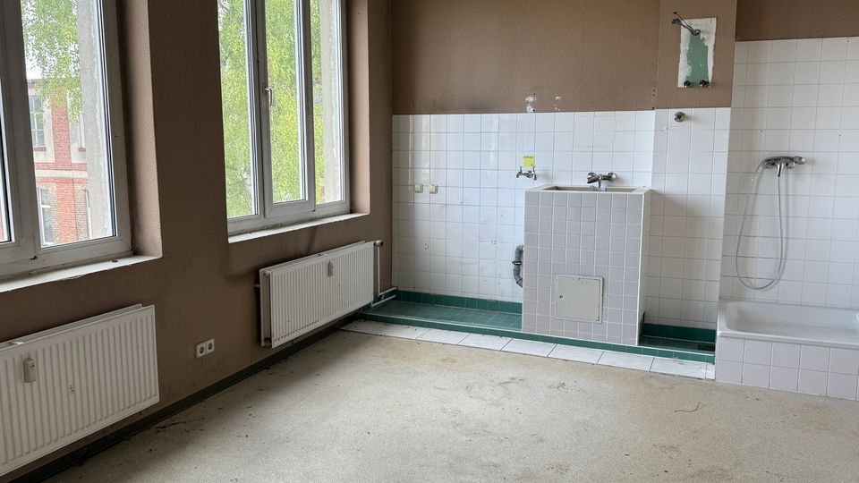 Leerer, teilweise gefliester Sanitärraum mit großen Fenstern, Heizkörpern und beigefarbenen Wänden. An der rechten Wand befinden sich ein gefliester Wasch-/Wasseranschlussbereich und eine Badewanne mit Duscharmatur; der Boden ist unbedeckt und wirkt unfertig.