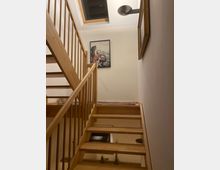 Holztreppe in einem Innenraum mit hellem Geländer, einem Dachfenster an der Decke und einem Bild an der Wand. Die Beleuchtung ist warm, und auf dem oberen Treppenabsatz liegt ein schmaler Teppich.