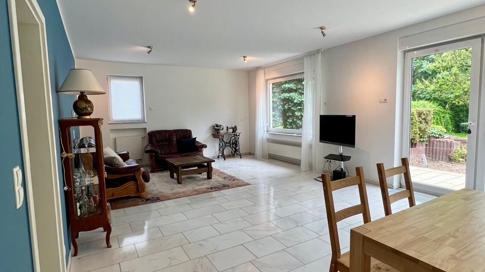 Großzügiger Wohn- und Essbereich mit hellen Fliesen, weißer Decke und Spotbeleuchtung, Sitzgruppe und Vitrine im hinteren Bereich sowie Esstisch auf rotem Teppich im Vordergrund. Große Fenster und eine Terrassentür lassen Blick ins Grüne; ein Fernseher steht an der rechten Wand.