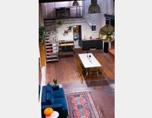 Industrieller Loft-Raum mit offener Gestaltung, Holzboden, einer Küchenzeile, einem Esstisch mit transparenten Stühlen, einer blauen Couch auf einem Orientteppich und einer Treppe, die zu einer kleinen Empore mit Schreibtisch führt. Eine große goldene Discokugel und industrielle Deckenlampen hängen von der Decke.