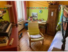 Ein bunt eingerichtetes Kinderzimmer mit Holzboden und grün-gelb gestrichenen Wänden, verziert mit einer Tier-Bordüre. Es enthält ein Bett, einen Holzschrank, einen Schreibtisch mit Utensilien, einen gelben Sessel, ein Keyboard auf einer roten Kommode und verschiedene dekorative Elemente.