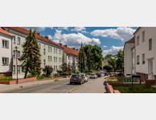 Wohnstraße mit mehrstöckigen Wohnhäusern, teils in Pastellfarben gestrichen, rote Ziegeldächer und gepflegte Vorgärten. Die Straße ist mit Autos gesäumt, daneben ein Gehweg; im Hintergrund sind Bäume und ein blau bewölkter Himmel sichtbar.