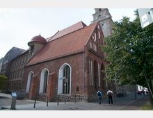 Backsteinkirche mit roten Ziegeldach und gotischen Spitzbogenfenstern, angelehnt an ein modernisiertes Gebäude, flankiert von einem Baum und Gehweg; der Hintergrund zeigt eine Turmspitze.