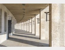 Überdachter Gang mit einer langen Reihe von Steinpfeilern, die rechteckige Lampen tragen. Die Wände und Pfeiler bestehen aus beigem Naturstein, und mehrere Schilder mit Standortinformationen befinden sich an den Seiten.