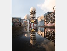 Ehemalige Abhörstation auf dem Teufelsberg in Berlin mit markanten Kuppeln und einer Fassade, die mit Graffiti und Wandgemälden bedeckt ist. Eine Pfütze im Vordergrund spiegelt die Gebäude und den Himmel wider.