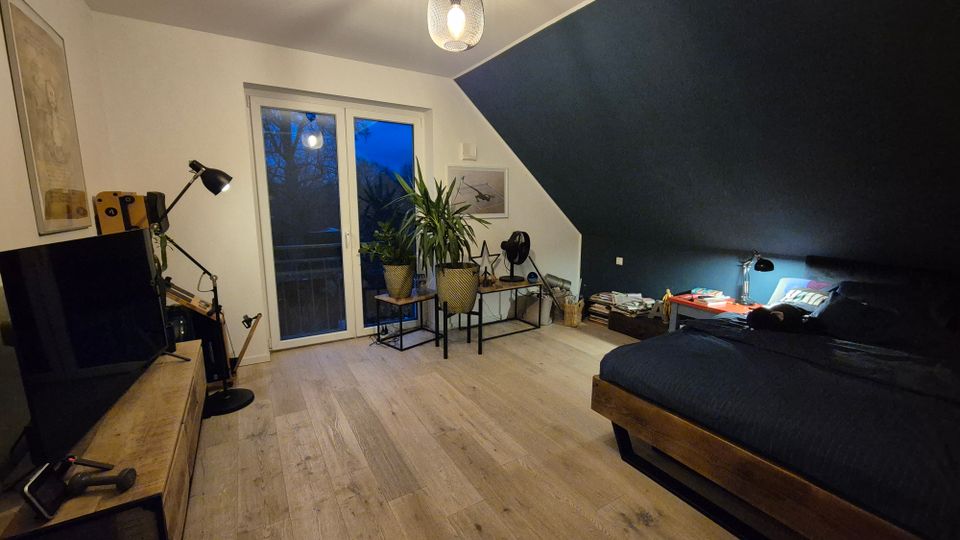 Schlafzimmer mit dunkler Akzentwand, Holzboden und minimalistischer Einrichtung. Ein Bett mit dunkler Bettwäsche steht rechts, links ein Fernseher auf einem Holztisch, daneben mehrere Pflanzen vor einer Balkontür.