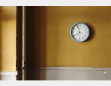 Eine gelb gestrichene Wand mit abgenutzter Oberfläche, an der eine runde Wanduhr mit blauem Rahmen hängt. Im linken Bildbereich befindet sich eine offene Tür mit einem weißen Türrahmen.