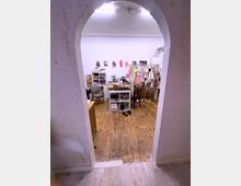 Ein Arbeitsraum mit Holzboden, aufgenommen durch einen bogenförmigen Durchgang. Im Raum befinden sich ein Schreibtisch, Regale mit Materialien, bunte Kleidungsstücke an einer Kleiderstange und Bilder an der weißen Wand.