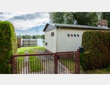 Ein kleiner Garten mit einem roten Metallzaun und dichtem Heckenschnitt grenzt an ein einfaches weißes Gebäude mit Flachdach und bunten Glasfenstern. Im Hintergrund sind ein See, eine Wiese sowie ein Strommast sichtbar.