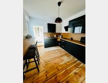 Moderne Wohnküche mit U-förmiger Einbauküche in mattschwarzen Fronten, Holzarbeitsplatte und Holzrückwand, dazu Holzdielenboden und schwarze Pendelleuchte. Links steht eine schmale Theke mit Hockern, hinten führt eine Tür in ein helles Bad mit sichtbarer Dusche.