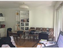 Ein Wohnzimmer mit Holzboden, einem weißen Regal voller Bücher und Dekorationsgegenstände, einem Tisch mit Giraffenfigur und einem Wäscheständer, auf dem Kleidung hängt. Rechts befindet sich eine große Fensterfront, die viel Tageslicht hereinlässt.