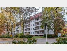 Mehrgeschossiges Wohnhaus mit weißer Fassade und roten Dächern, umgeben von herbstlichen Bäumen und Sträuchern. Im Vordergrund Kreuzung mit Straßenschildern „Westfalenring“ und „Gronauer Weg“.