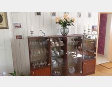 Wohnzimmerbereich mit einer langen Glasvitrine aus Holz, gefüllt mit Gläsern und Porzellan, vor einer weiß vertäfelten Wand mit mehreren gerahmten Fotos. Auf der Vitrine stehen Kerzenhalter, ein Herzdeko-Element und eine große Vase mit Blumen; rechts führt eine offene Tür in einen Flur mit gemusterter Tapete.
