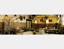 Gemütlicher Gastraum mit Holzmöbeln und grün gedeckten Tischen. An den Wänden hängen ausgestopfte Tiere, Bilder und rustikale Dekorationen; im Hintergrund ein großer Kachelofen und eine geschlossene Tür.