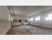 Ein leerer Raum mit abgenutztem Boden, bröckelnden weißen Wänden und großen Fenstern, die natürliches Licht hereinlassen. Im hinteren Bereich befinden sich Trennwände mit Türen und Fensterausschnitten; Heizkörper sind entlang der rechten Wand angebracht.