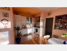 Helle Küche in einer Wohnung mit cremefarbenen Wänden und oranger Decke, ausgestattet mit weißen Schränken, einer Arbeitsplatte aus Holz und einem Herd. Der Raum ist dekoriert mit bunten Gemälden, Pflanzen und einer Tiger-förmigen Teppichmatte auf dem Fliesenboden.