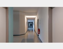 Langer, gerader Flur in einem Gebäude mit hellbeigen Wänden, blau-grauem Boden und Deckenbeleuchtung. An den Wänden hängen Bilderrahmen, rechts hängt ein Feuerlöscher, und am Ende des Flurs ist eine Glastür mit einem Notausgangszeichen sichtbar.