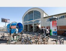 Eingangsbereich des U-Bahnhofs Warschauer Straße in Berlin mit einem historischen Gebäude aus Glas und Metall. Im Vordergrund befinden sich zahlreiche abgestellte Fahrräder, eine blaue Telefonzelle und eine Litfaßsäule mit Werbeplakaten.