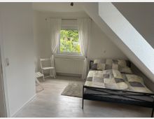 Ein schlicht eingerichtetes Schlafzimmer mit schräger Decke, einem Metallbett mit bunter Bettwäsche, einem großen Fenster mit weißen Vorhängen und hellem Laminatboden. Neben dem Fenster stehen zwei weiße Klappstühle, und ein kleiner Teppich liegt vor dem Bett.