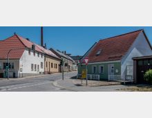 Straßenecke in einer Kleinstadt mit Wohnhäusern und roten Ziegeldächern. Im Hintergrund ist ein Fabrikschornstein zu sehen, und eine Verkehrsampel sowie Straßenschilder stehen im Vordergrund.