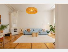 Wohnzimmer mit heller Einrichtung, einer blauen Couch, einem Glastisch und einem dekorativen Kaminofen mit weißen Verzierungen. Der Raum hat einen Holzboden mit geometrischem Muster, Stuckdecken und ist mit Pflanzen und einer modernen Hängelampe ausgestattet.