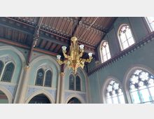 Innenansicht einer Kirche mit gotischen Elementen, darunter hohe Spitzbogenfenster mit Buntglas und reich verzierten Rahmen. Von der Holzdecke hängt ein goldener Kronleuchter mit kunstvollen Details.