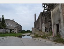 Verlassene Industrieanlage mit großen, grauen Gebäuden aus Beton, teils beschädigten Fassaden und offenen Strukturen. Eine Schornsteinstruktur ragt in die Höhe, während im Vordergrund Betonflächen und eine Pfütze sichtbar sind, umgeben von überwuchertem Grün.