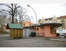Kleiner Platz mit einem runden, rosafarbenen Gebäude mit flachem, auskragendem Dach und Holztresen; daneben ein kleiner, hölzerner Schuppen mit grünem Wellblechdach. Im Hintergrund stehen Altbauten mit verzierten Fassaden und ein moderner, orangefarbener Wohnblock.