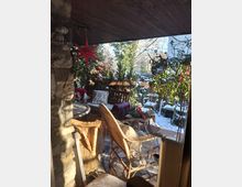 Überdachte Terrasse mit Natursteinwand und Holzdecke, eingerichtet mit Korbmöbeln und winterlicher Dekoration. Im Hintergrund sind schneebedeckte Pflanzen und Bäume sichtbar.