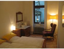 Schlafzimmer mit Holzboden und einem Doppelbett mit weißen Bettdecken und gelben Kissen. Im Raum befinden sich ein antiker Waschtisch mit Marmorplatte, ein roter Polstersessel, eine Stehlampe sowie ein Fenster mit Blick auf eine Außenfassade.