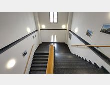 Treppenhaus mit dunklen Stufen und hellen Wänden, beleuchtet durch runde Wandlampen. An den Wänden hängen Bilder, und zwei Fenster lassen natürliches Licht einfallen.