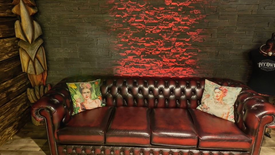 Innenraum mit dunkelrotem, gepolstertem Chesterfield-Ledersofa vor einer grauen Steinwand, die mittig von rotem Akzentlicht beleuchtet wird. Holzboden, dekorative Kissen, eine geschnitzte Holzskulptur links und ein niedriger Tisch mit roten Fächern im Vordergrund.