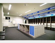 Umkleideraum einer Sportmannschaft mit offenen Spinden, einer langen weißen Arbeitsinsel in der Mitte und Sitzbänken entlang der Wände. An der Decke hängen blaue Banner mit Begriffen wie „Vertrauen“ und „Siegeswille“.