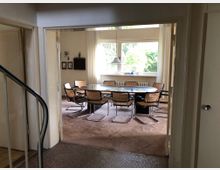 Ein heller Esszimmerbereich mit einem ovalen Glastisch, der von mehreren Stühlen mit geflochtenen Sitzflächen und Metallrahmen umgeben ist. Große Fenster lassen viel Tageslicht herein, mit Blick auf grüne Vegetation draußen; im Hintergrund stehen eine Heizung und dekorative Wandelemente.