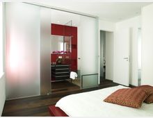 Modernes Schlafzimmer mit dunklem Holzboden, weißer Bettwäsche und roten Dekokissen. Eine halbtransparente Schiebetür trennt den Schlafbereich von einem angrenzenden Badezimmer mit roten Wänden, schwarzer Waschtischmöbel und dekorativen Orchideen.