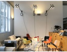 Ein moderner Raum mit einer weißen Wand, an der drei verstellbare Wandleuchten angebracht sind, sowie einer Sitzgruppe aus verschiedenen Stühlen und Sofas in unterschiedlichen Farben und Designs. Auf der linken Seite befindet sich ein großes Fenster mit Jalousien, während rechts Holzmöbel und gestapelte Gegenstände erkennbar sind.