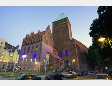 Backsteingotische Kirche mit spitz zulaufendem Turm und grünem Kupferdach, beleuchtet bei Dämmerung. Im Vordergrund geparkte Autos, Fahrräder und Banner an einem modernen Gebäude links, rechts säumen Bäume die Straße.