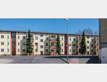 Mehrstöckiges Wohngebäude mit cremefarbenen Fassaden und roten Backsteinverzierungen, mehreren Balkonen und Giebeldächern. Vor dem Gebäude befindet sich eine gepflasterte Straße mit Verkehrszeichen, umgeben von einzelnen Bäumen und Laternen.