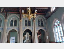 Innenansicht eines historischen Kirchenraums mit spitzbogigen Fenstern und transparenter Glasmalerei. Im Vordergrund hängt ein großer, goldener Kronleuchter, darunter ein weißes Orgelgehäuse mit goldenen Verzierungen, neben einer geschwungenen Holztreppe.