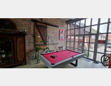 Industrie-inspirierter Innenraum mit Backsteinwand und einer großen Glasfront, die den Blick nach draußen freigibt. Im Vordergrund steht ein Billardtisch mit pinkem Filz, daneben eine grüne Leiter und weitere dekorative Elemente wie ein Holzschrank und Wandbilder.