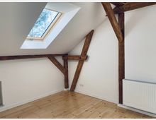 Zimmer unter einer Dachschräge mit weiß gestrichenen Wänden, sichtbaren Holzbalken, einem Oberlichter-Fenster und einem hellen Holzfußboden. Rechts ist eine weiße Heizkörperheizung zu sehen.