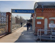 Eingang zum U-Bahnhof Warschauer Straße in Berlin, mit einem blauen Schild und den U-Bahn-Linien U1 und U3. Der Bahnhof besteht aus Backsteinarchitektur, einem teilweise überdachten Bahnsteig und umgebenden modernen Gebäuden im Hintergrund.