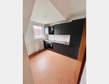 Helle, leere Küche mit schräger Decke, großem Fenster und Heizkörper unter dem Fenster. Entlang einer Wand verläuft eine moderne, dunkelgraue Einbauküche mit Herd, Dunstabzug, Spüle und weißem Fliesenspiegel.