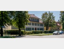 Straßenansicht mit Wohnhäusern im traditionellen Baustil, gekennzeichnet durch helle Fassaden, grüne Fensterläden und Satteldächer, flankiert von Bäumen und Hecken. Im Vordergrund verläuft eine Straße mit parkenden Autos und einem Gehweg.