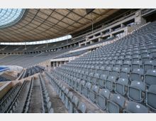 Innenansicht eines großen Stadions mit mehreren Sitzreihen aus grauen Kunststoffsitzen. Die Tribünen sind überdacht, und das Dach besteht aus einer transparenten, weitgespannten Konstruktion.