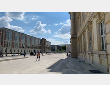 Weitläufiger Platz mit gepflastertem Boden, flankiert von historischen und modernen Gebäuden mit großen Fenstern; rechts ein helles, detailreich verziertes Gebäude, links ein moderner Bau mit roten und weißen Fassadenelementen. Im Hintergrund führt ein Torbogen zu grünen Bäumen.