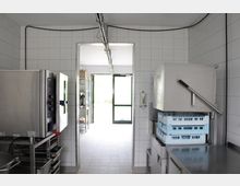 Industrieküche mit gefliesten Wänden und Boden, großen Edelstahlgeräten wie einem Kombidämpfer und einer Spülmaschine mit blauen Geschirrkörben. Durch eine offene Tür ist ein angrenzender Raum sowie eine Glas-Eingangstür zu sehen, die nach draußen führt.