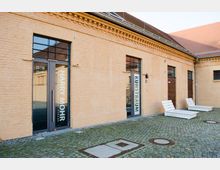 Ehemaliges Backsteingebäude mit beigefarbener Fassade und dunkelgrauen Metalltüren, beschriftet mit "Harry Mohr" und "Kunstraum". Der gepflasterte Vorplatz enthält weiße, moderne Sitzgelegenheiten.